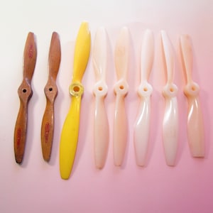 May include: A collection of eight model airplane propellers. The propellers vary in color, including wood-toned, yellow, and off-white. The propellers are arranged on a pink background.