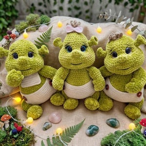 Puede incluir: Tres muñecos verdes de ganchillo hechos a mano con ojos grandes y caras sonrientes. Cada muñeco lleva un pañal blanco y tiene orejas pequeñas. Un muñeco tiene un lazo rosa y otro un accesorio para el pelo marrón. Los muñecos están colocados sobre una superficie de color neutro con musgo decorativo y luces de hadas.