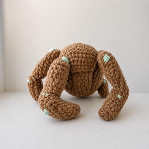 May include: A brown crocheted creature with a rounded central body and four thick, segmented limbs. The yarn texture is consistent throughout, featuring small, light mint green accents stitched onto the joints and limbs of the figure.