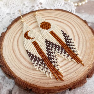 Op de afbeelding: Een paar lange, geperst oorringen met een geometrisch patroon in bruin, wit en goud. De oorringen hangen aan een houten oppervlak.