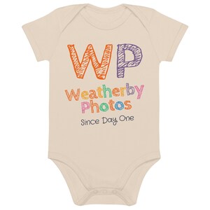 May include: A beige baby onesie with short sleeves. The onesie has the text "WP Weatherby Photos Since Day One" in colorful, handwritten-style lettering. The letters "WP" are in orange and purple.