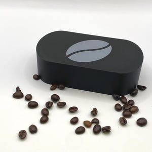 Op de afbeelding: Een zwarte, rechthoekige container met een grijs koffieboonontwerp erop. Verspreid rond de container liggen talrijke geroosterde koffiebonen, die een product met koffiethema laten zien.