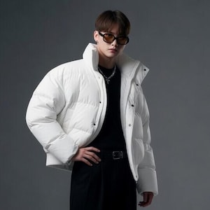 May include: A white puffer jacket with a high collar, worn over a black long-sleeved shirt and black trousers. The outfit includes black lace-up boots and dark-tinted sunglasses. The jacket features horizontal quilted panels and a front zipper closure.