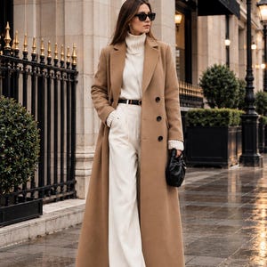 Puede incluir: Un abrigo largo de lana color camel sobre un suéter de cuello alto blanco acanalado y pantalones blancos de talle alto y pierna ancha. El conjunto incluye un cinturón negro fino con hebilla dorada, gafas de sol negras y un pequeño bolso de mano negro.