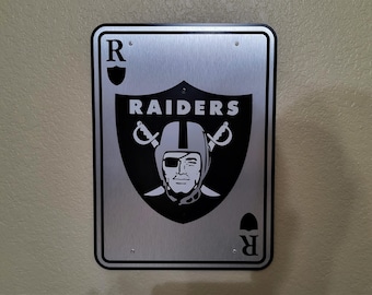 Puede incluir: Un letrero metálico rectangular con el logotipo de los Raiders de Las Vegas. El letrero tiene un fondo plateado cepillado con el logotipo del equipo en blanco y negro. El logotipo incluye una cara con casco, espadas cruzadas y la palabra "RAIDERS". Las esquinas del letrero son redondeadas.