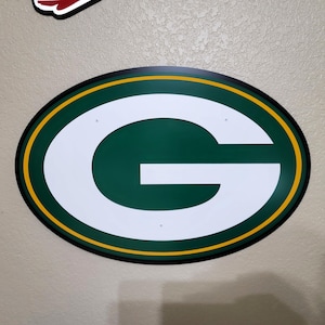 May include: An oval-shaped sign featuring the Green Bay Packers logo. The logo is a white letter "G" on a green background, outlined with a yellow and black border. The sign is mounted on a light-colored wall.