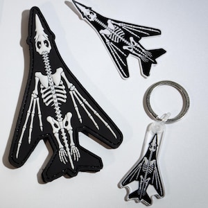 May include: A rectangular brown card with black airplane illustrations and the text Gunship Garage Company Thank You For Supporting Our Small Business, a large black patch, a small black pin, and a black keychain, all featuring white skeleton designs.