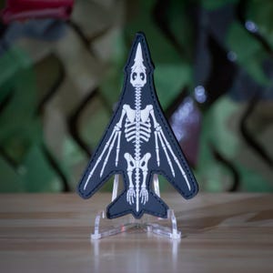 May include: A black patch shaped like a fighter jet with a white skeletal print of a bird or bat-like creature. The patch features a detailed spine, rib cage, and wing bone structure, with stitched edges and a pointed nose and tail design.