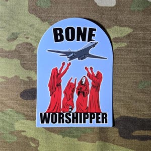 May include: A sticker with a light blue background featuring a B-1 bomber and figures in red robes with outstretched arms. The words "BONE WORSHIPPER" are printed in black.