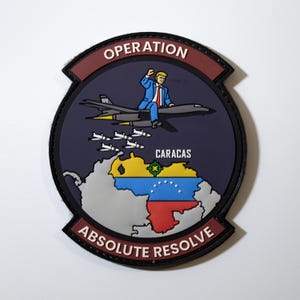 May include: A circular morale patch featuring a dark blue background with the text OPERATION at the top and ABSOLUTE RESOLVE at the bottom. It depicts a figure in a blue suit riding a gray bomber aircraft dropping missiles toward a map of Venezuela.