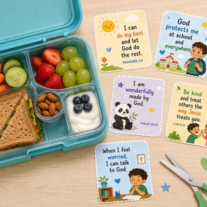 May include: A light blue bento box filled with a sandwich, sliced cucumbers, strawberries, grapes, almonds, and yogurt, accompanied by five square cards with religious text and illustrations, and a pair of green-handled metal scissors.