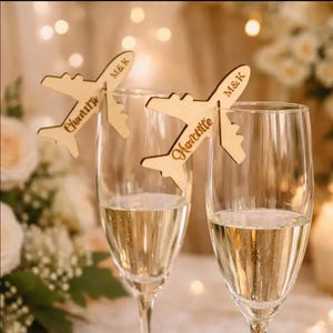 Peut inclure: Deux flûtes à champagne avec des marque-places en forme d'avion en bois. Les marque-places sont gravés de noms et d'initiales. Les flûtes sont remplies d'une boisson pétillante, et l'arrière-plan est flou avec des lumières et des fleurs.