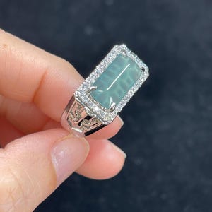 Könnte beinhalten: Ein silberner Ring mit einem rechteckigen, hellgrünen Edelstein. Der Edelstein ist von kleinen, klaren Steinen umgeben. Der Ring hat ein verziertes, durchbrochenes Design an den Seiten.