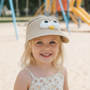 May include: A straw visor with a whimsical penguin design. The visor features large white eyes, a yellow beak, and a light brown brim. The child is wearing a floral print dress. The background includes playground equipment.