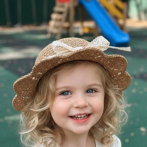 May include: A straw hat with a scalloped brim and a white lace bow. The hat is a light brown color and appears to be made of woven straw. The background shows a playground with a blue slide.