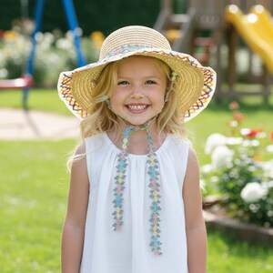 May include: A young girl wearing a straw sun hat with a colourful embroidered brim and a white dress. The hat has a floral patterned ribbon tie. The background shows a playground and green grass.