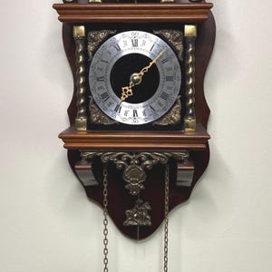Könnte beinhalten: Antike Wanduhr mit dunklem Holzrahmen und aufwendigen Messingakzenten. Das Zifferblatt hat römische Ziffern und dekorative Zeiger. Ein Pendel mit zwei Messinggewichten hängt darunter. Die Oberseite zeigt eine Skulptur und den Text "CONU ELCK GOSYN SINE".
