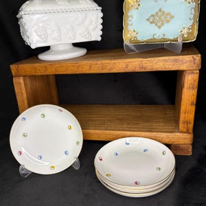 Può includere: Una bomboniera bianca con coperchio e motivo a uva, un vassoio rettangolare blu e oro con accenti floreali e diversi piccoli piatti bianchi con motivi a pois colorati. Gli oggetti sono esposti su una mensola di legno.