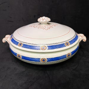 Puede incluir: Un plato de cerámica blanco con tapa. El plato presenta un patrón floral azul y naranja alrededor del borde y en la tapa. La tapa tiene un asa decorativa y una línea marrón.