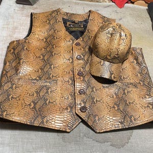 May include: A sleeveless vest and a baseball cap, both featuring a tan and brown snakeskin pattern. The vest has a V-neckline, five dark brown buttons, and two front pockets. A black label inside the vest reads LUXURIOUS LEATHER WEAR.