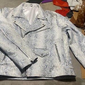 May include: A motorcycle jacket featuring a white and grey snakeskin pattern, a notched lapel, silver-toned metal zippers on the front and sleeves, a small snap-button pocket, and a smooth white interior lining visible at the collar area.