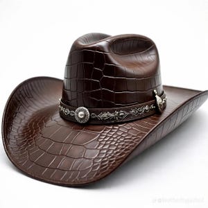 May include: A brown cowboy hat with a textured, crocodile-like pattern. The hat features a decorative silver band with floral and circular accents. The hat's brim is wide, and the crown is slightly curved.