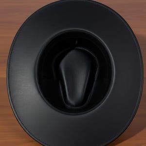 Pu&ograve; includere: Un cappello fedora nero con tesa larga. Il cappello &egrave; realizzato in un materiale liscio simile alla pelle. L'interno del cappello &egrave; anch'esso nero e ha una corona arrotondata. Il cappello &egrave; appoggiato su una superficie di legno.