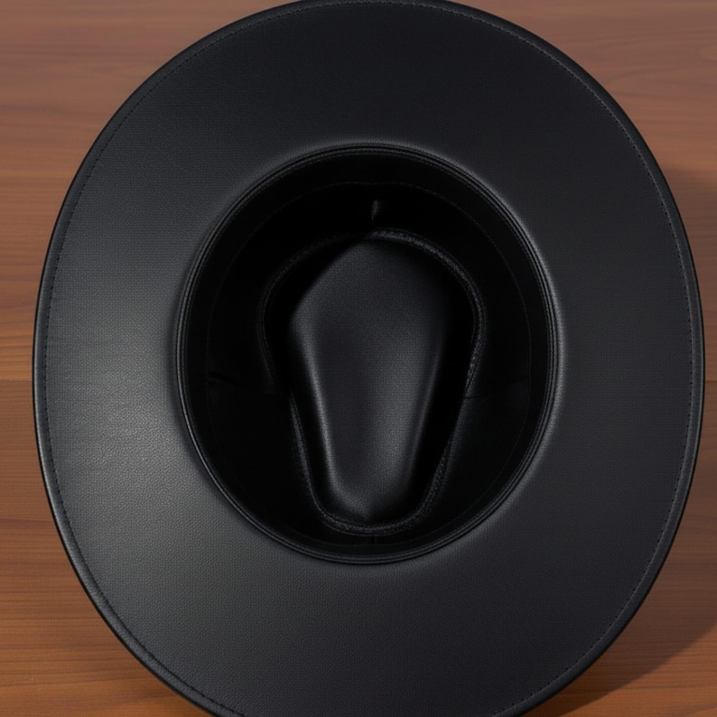 Pu&ograve; includere: Un cappello fedora nero con tesa larga. Il cappello &egrave; realizzato in un materiale liscio simile alla pelle. L'interno del cappello &egrave; anch'esso nero e ha una corona arrotondata. Il cappello &egrave; appoggiato su una superficie di legno.