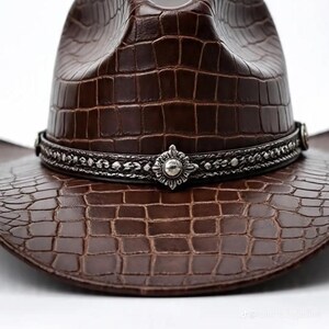 May include: A brown cowboy hat with a textured, crocodile-like pattern. The hat features a decorative silver band with an ornate centerpiece. The hat's brim and crown are visible against a white background.