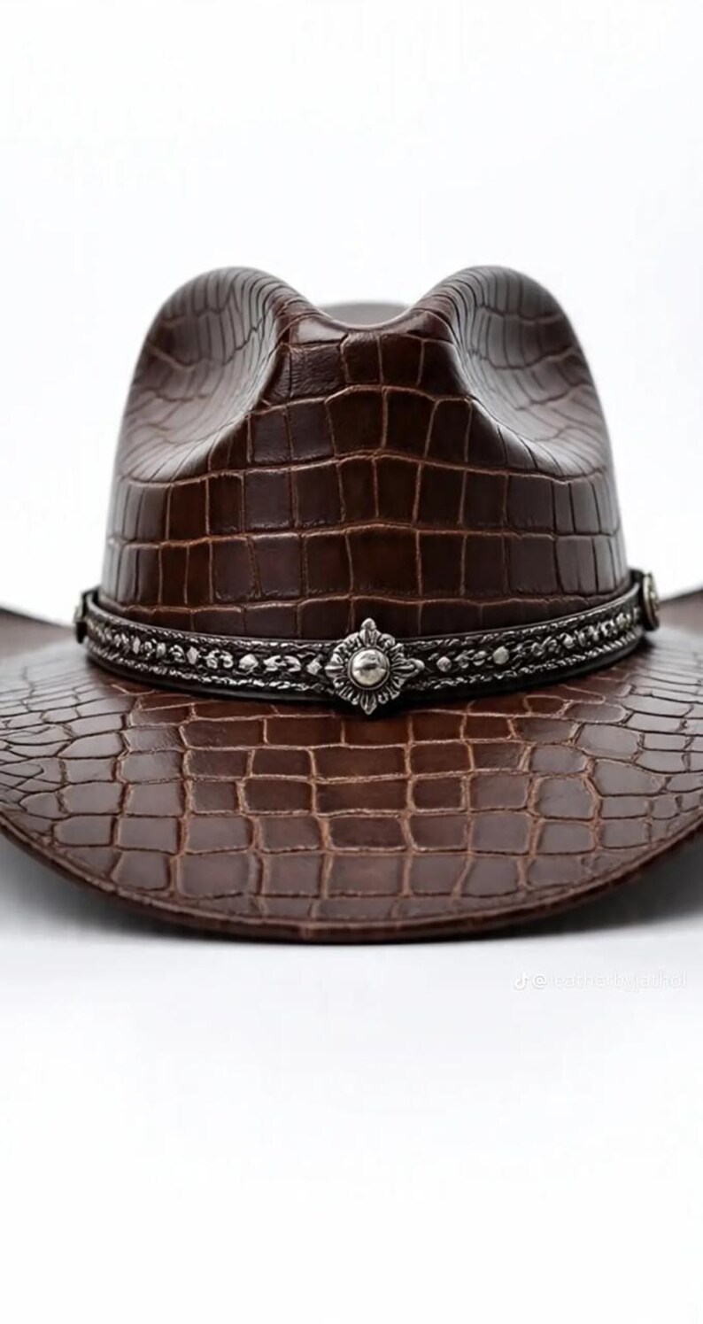 May include: A brown cowboy hat with a textured, crocodile-like pattern. The hat features a decorative silver band with an ornate centerpiece. The hat's brim and crown are visible against a white background.