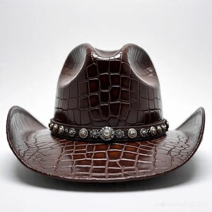 May include: A brown cowboy hat with a textured, crocodile-like pattern. The hat features a decorative silver band with circular embellishments around the crown. The hat has a classic cowboy hat shape with a curved brim.