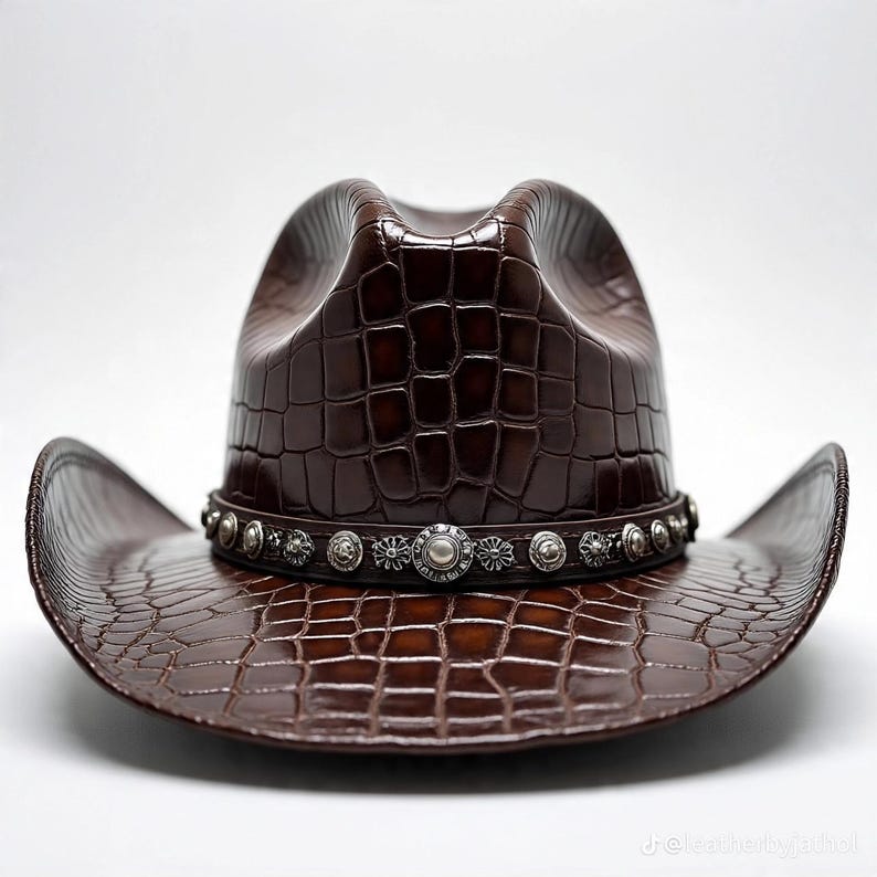 May include: A brown cowboy hat with a textured, crocodile-like pattern. The hat features a decorative silver band with circular embellishments around the crown. The hat has a classic cowboy hat shape with a curved brim.