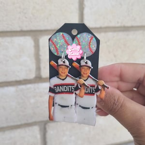 May include: Baseball-themed earrings featuring two baseballs with red stitching and glitter accents. Below, two figures in white and black baseball uniforms with the word "Bandits" are visible. The earrings are attached to a black tag.