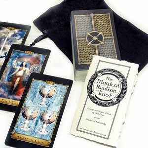 May include: A black velvet bag containing a deck of tarot cards featuring the Magical Realism Tarot title. The cards have black backs with gold accents. Three cards are laid out on a white surface: The World, Ace of Swords, and Three of Cups.