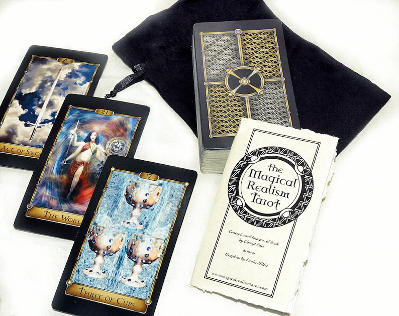 May include: A black velvet bag containing a deck of tarot cards featuring the Magical Realism Tarot title. The cards have black backs with gold accents. Three cards are laid out on a white surface: The World, Ace of Swords, and Three of Cups.