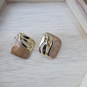 May include: A pair of square earrings with a gold-colored wave design on a light brown background. The earrings are displayed on a white wooden hexagonal tray with a jute rope handle, set against a wooden surface.