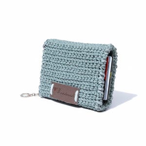 May include: A crocheted wallet in a muted teal color. The wallet has a rectangular shape with a flap closure. A small brown leather tag with the word "Handmade" is attached. A silver-colored key ring is attached to the side.