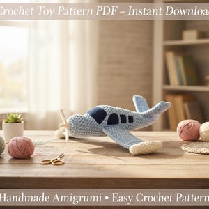 Op de afbeelding: Een lichtblauw gehaakt vliegtuig met donkerblauwe accenten en een houten propeller ligt op een houten tafel. De afbeelding toont bollen garen in roze, groen en crème, een kleine plant en de tekst "Crochet Toy Pattern PDF - Instant Download".