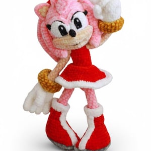 Peut inclure: Poupée Amy Rose au crochet faite à la main, avec une tête et des piquants roses, une robe rouge avec une bordure blanche et des chaussures rouges. La poupée lève deux doigts. Le texte "Crochet Toy Pattern PDF - Instant Download" et "Handmade Amigurumi • Easy Crochet Pattern" sont visibles.