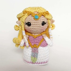 Peut inclure: Poupée au crochet faite à la main représentant un personnage avec des cheveux blonds, une couronne dorée et une robe rose et blanche. La poupée a de grands yeux noirs et mesure environ 15 cm de haut. Objet artisanal unique.