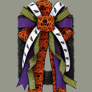 May include: A decorative wreath featuring a large bow made of multiple ribbons. The ribbons display patterns of black skulls on orange fabric, black crows on white fabric, and alternating purple and lime green stripes on a black textured base.