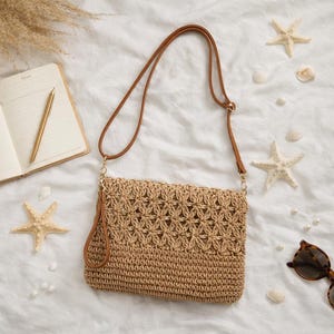 Puede incluir: Bolso bandolera beige tejido con correa y asa de cuero marrón. El bolso presenta un estampado floral y se muestra sobre una superficie blanca con estrellas de mar, conchas, un cuaderno, un bolígrafo y gafas de sol.