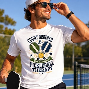 May include: White t-shirt with a graphic design featuring pickleball paddles, a ball, and the text "Court Ordered Pickleball Therapy." The design includes stars and a banner that reads "Dink Responsibly."