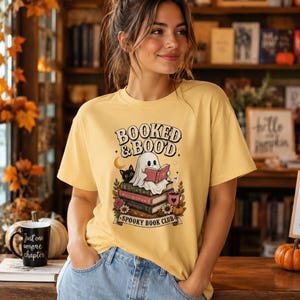 Peut inclure: T-shirt jaune avec l'inscription "Booked & Boood" et un dessin d'un fantôme lisant un livre, un chat noir et "Spooky Book Club". La personne porte le t-shirt avec les mains dans les poches.