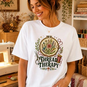 Può includere: T-shirt bianca con un ricamo colorato con fiori, forbici e una spoletta di filo. Il testo "THREAD THERAPY" è visualizzato sotto il ricamo. La persona che indossa la maglietta sta sorridendo.