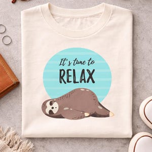 Peut inclure: T-shirt crème avec une illustration de paresseux de dessin animé. Le paresseux est brun et allongé devant un cercle bleu avec le texte "It's time to RELAX". Le t-shirt est plié sur une surface grise.