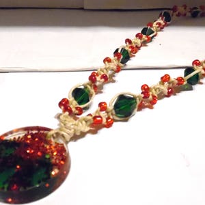 May include: A handmade necklace featuring a pendant with red glitter and green accents. The necklace has a natural cord with red and green beads. The pendant is oval-shaped and the beads are small and round.