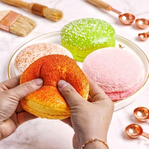May include: An orange, textured pastry being squeezed by hands, with a plate of colorful, frosted pastries in the background. Other pastries are green, pink, and peach. Copper measuring spoons and a pastry brush are also visible.