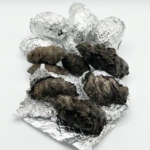May include: A collection of dark, organic-looking objects, some wrapped in shiny silver foil. The objects vary in size and shape, with a mix of dark brown and black hues. The foil adds a contrasting metallic element to the composition.