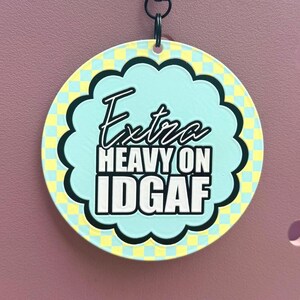 Pode incluir: Um porta-chaves circular com um centro recortado azul claro contendo o texto a preto e branco Extra HEAVY ON IDGAF. O fundo apresenta um padrão de xadrez amarelo e azul claro. Está suspenso por um fecho de metal preto e um cordão preto.